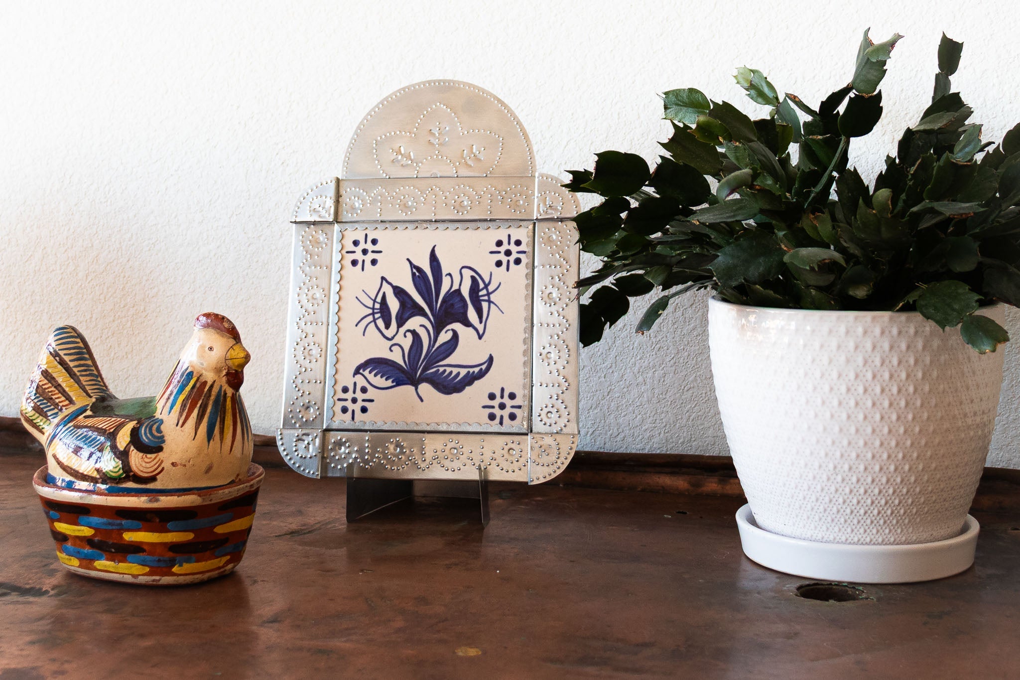 A handcrafted tinwork frame featuring a blue and white hand-painted tile, displayed on a copper surface. A ceramic rooster dish and a potted cactus sit nearby, with a white wall in the background.